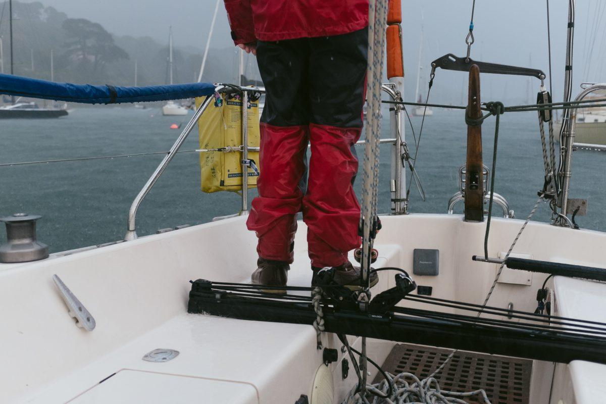 A sailor is standing on the deck of a boat. It is raining. The boat is in the Falmouth estuary, Cornwall.